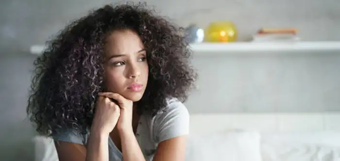 A sad young female with curly hair looking off into the distance with her head on her hands (700x333 vertical tab)