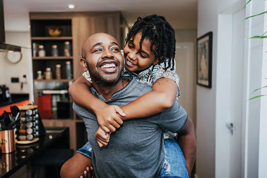 A father giving his young daughter a piggy back ride in their kitchen